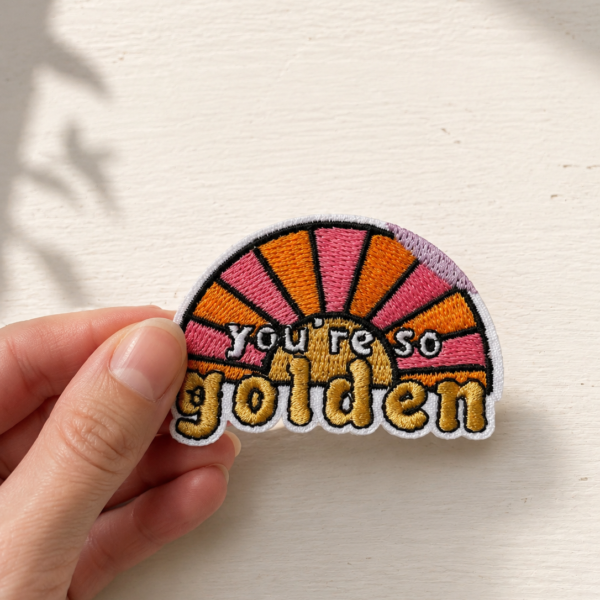 Broderet strygemærke med teksten "You're so golden" foran en rosa og orange solopgang. Mærket holdes mellem fingrene foran en lys baggrund.