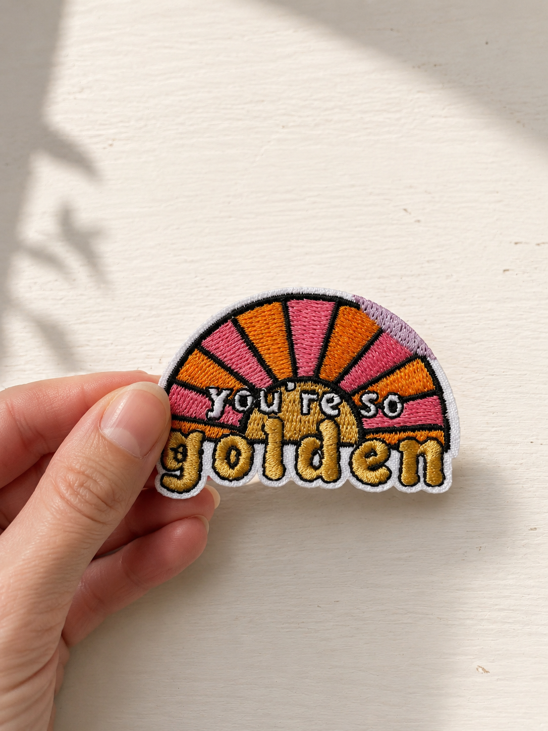 Broderet strygemærke med teksten "You're so golden" foran en rosa og orange solopgang. Mærket holdes mellem fingrene foran en lys baggrund.