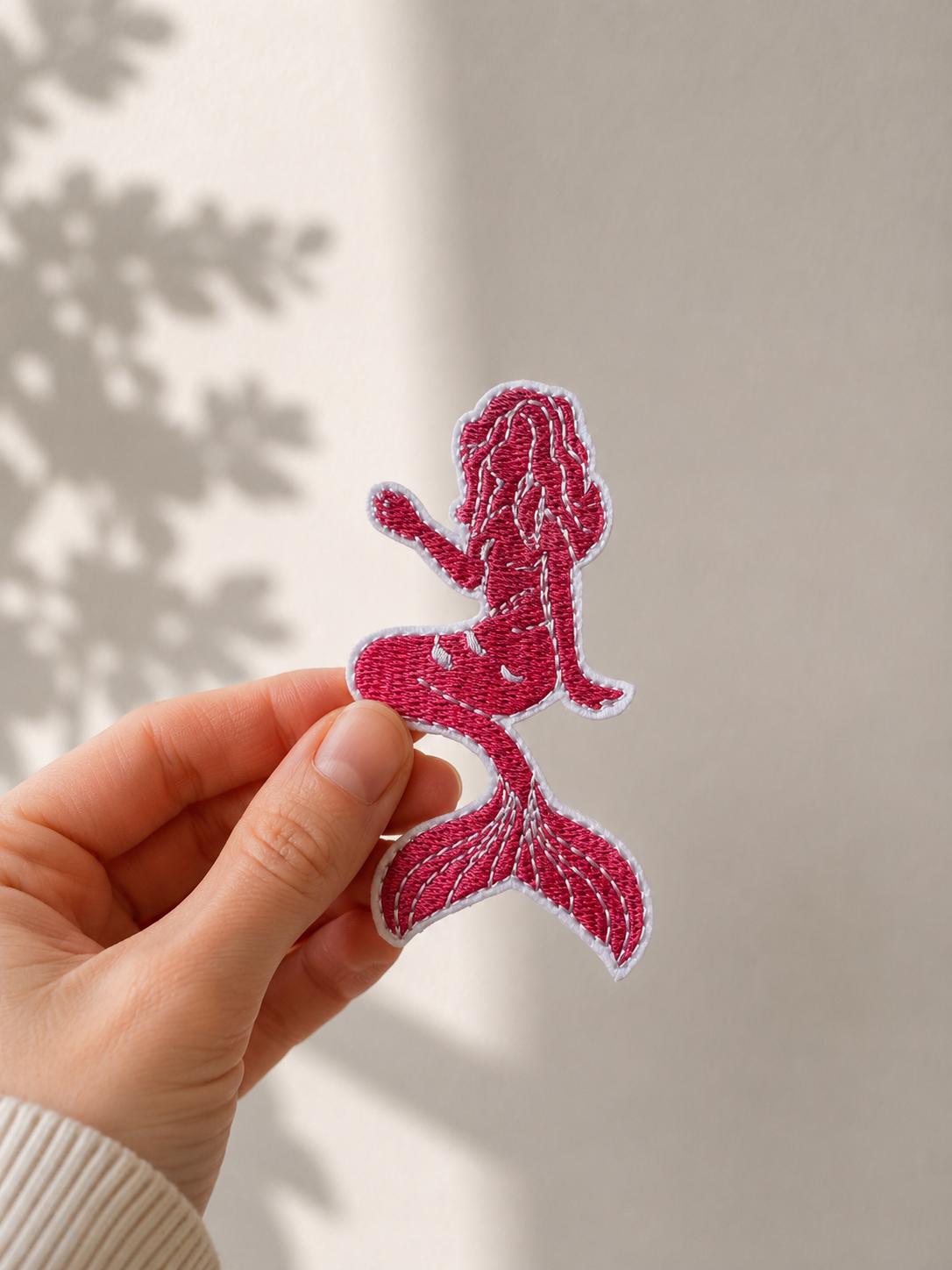 Broderet strygemærke formet som en pink havfrue med lang hale og bølgende hår. Mærket holdes mellem fingrene foran en lys baggrund.