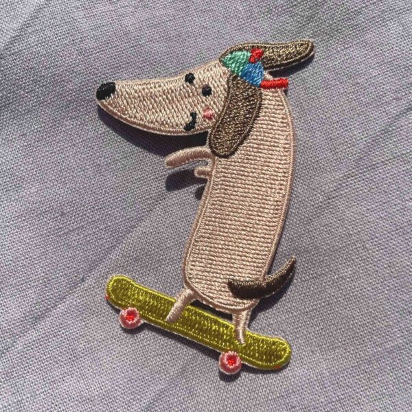 Broderet strygemærke med en gravhund på skateboard iført en farverig kasket. Perfekt til at reparere huller eller dekorere jakker, jeans, børnetøj, tasker og accessories.
