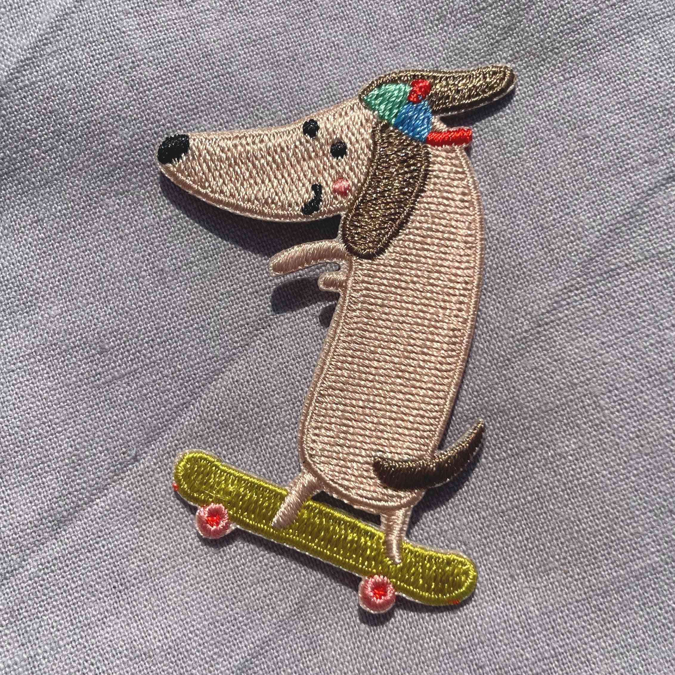 Broderet strygemærke med en gravhund på skateboard iført en farverig kasket. Perfekt til at reparere huller eller dekorere jakker, jeans, børnetøj, tasker og accessories.