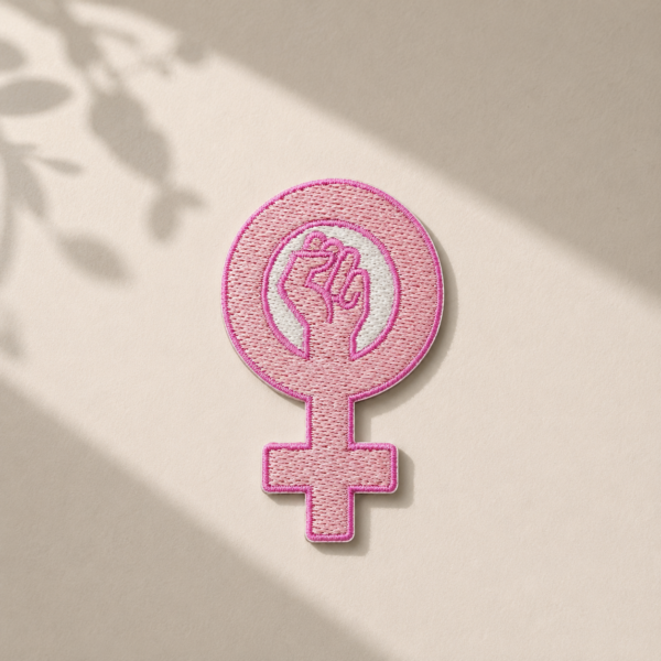 Broderet feminist strygemærke med lyserødt kvindetegn og knyttet næve på lys baggrund med naturligt lys. Iron-on patch til jakker, tasker, jeans og andet tøj.